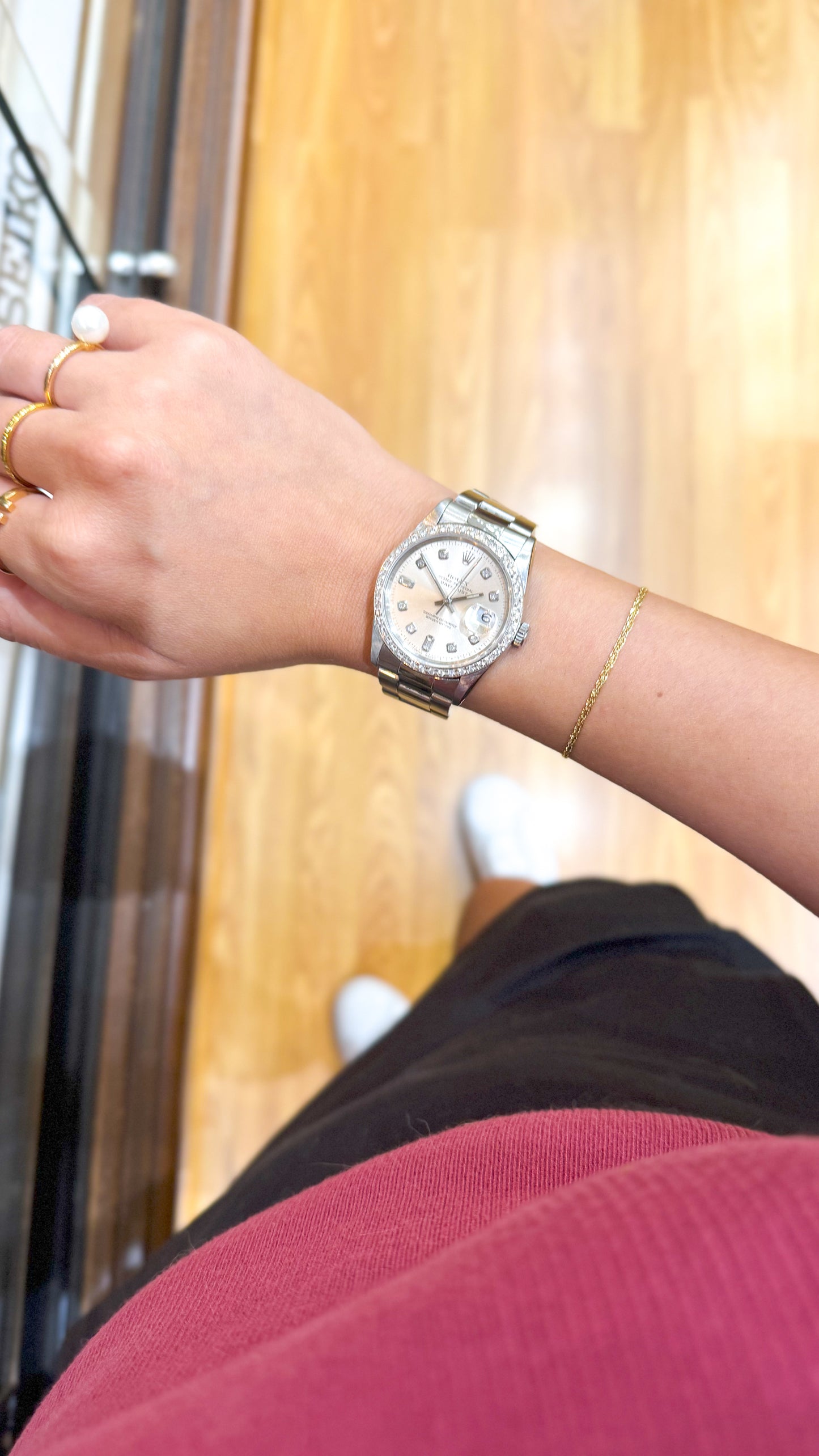 Rolex Datejust with diamond bezel worn on wrist, shown with gold jewelry over wood floor.